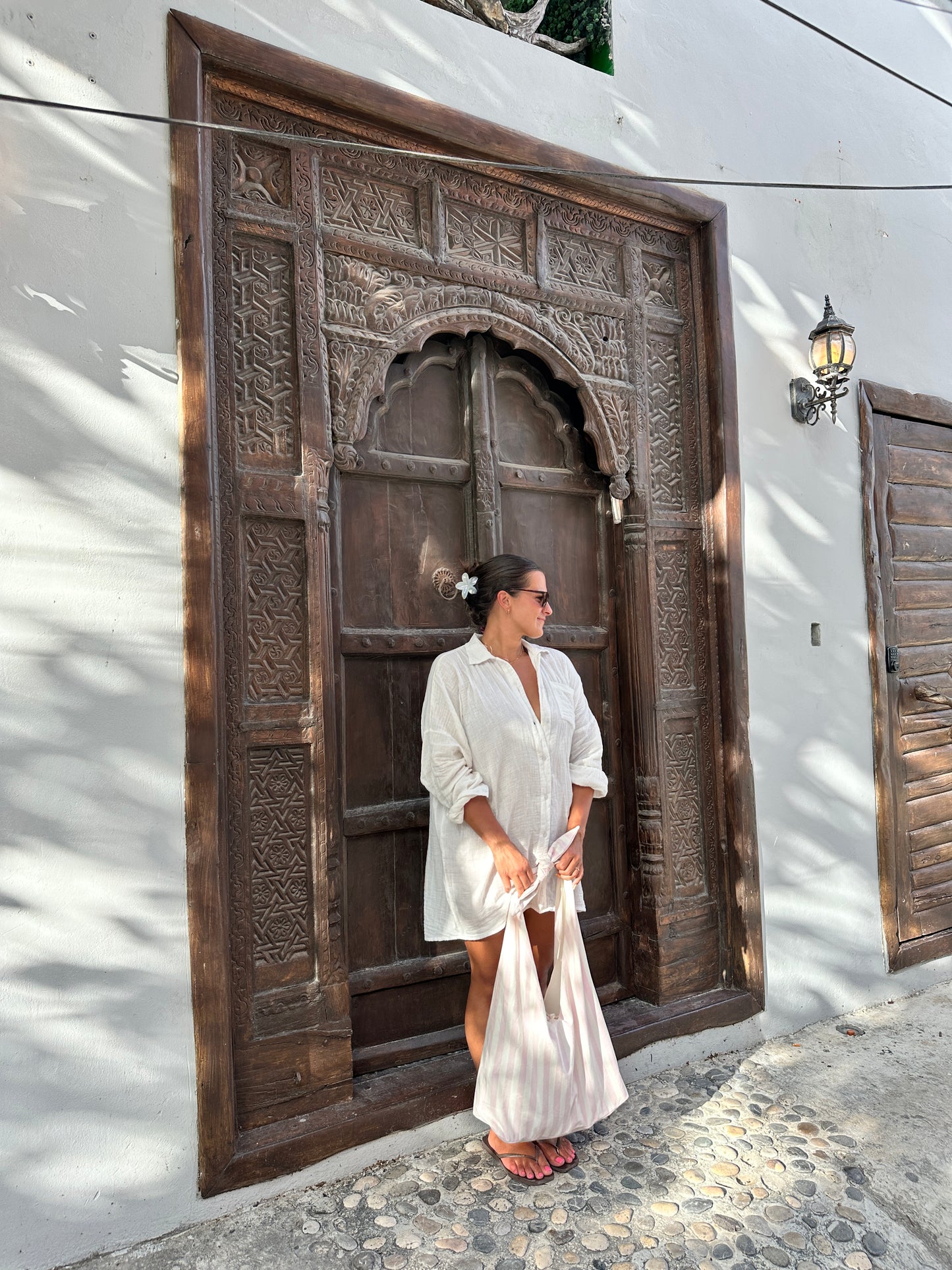 Woman in a white dress standing in front of a large wooden door with intricate carvings holding a pink and white beach bag