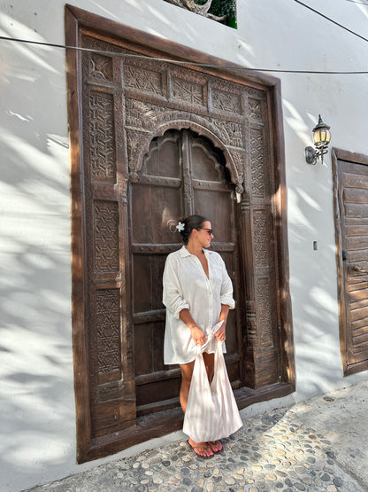 Woman in a white dress standing in front of a large wooden door with intricate carvings holding a pink and white beach bag